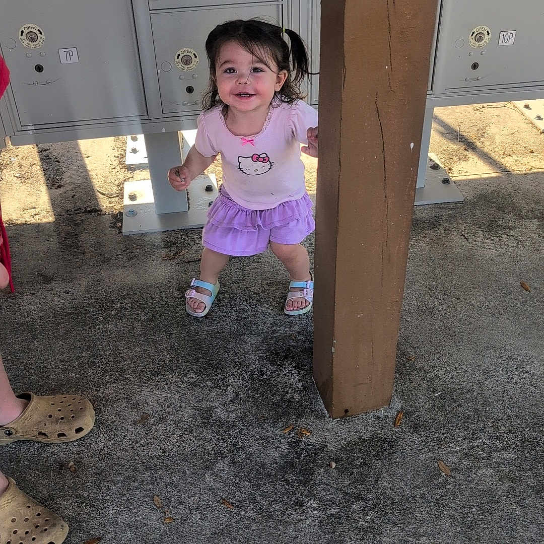 Bella is registered to the contest to win money with this photo: toddler, child, girl, smiling, hello_kitty, purple_skirt, sandals, post, mailboxes, concrete, leaves, shadow, outdoor, playful, person, clothing, footwear, hair, happy, standing