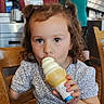 child, ice_cream, indoor, curly_hair, pigtails, wooden_chair, vanilla, cone, person, casual_clothing, cafe, background, food, eating, happy, girl, seated, table, light, smiling