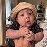 Gabriel is registered to the contest to win money with this photo: baby, infant, child, portrait, close_up, wide_eyes, hat, beige_hat, black_clothing, sitting, held, adult, hands, feet, indoor, home, living_room, shelf, couch, candid