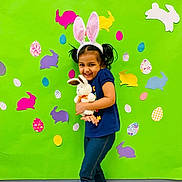 Dhriti is registered to the contest to win money with this photo: child, girl, bunny_ears, plush_toy, stuffed_animal, green_background, colorful, easter_eggs, paper_bunnies, smile, happy, cute, playful, indoor, casual_clothing, blue_shirt, leggings, purple_shoes, pigtails, holiday_theme