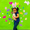 child, girl, bunny_ears, plush_toy, stuffed_animal, green_background, colorful, easter_eggs, paper_bunnies, smile, happy, cute, playful, indoor, casual_clothing, blue_shirt, leggings, purple_shoes, pigtails, holiday_theme
