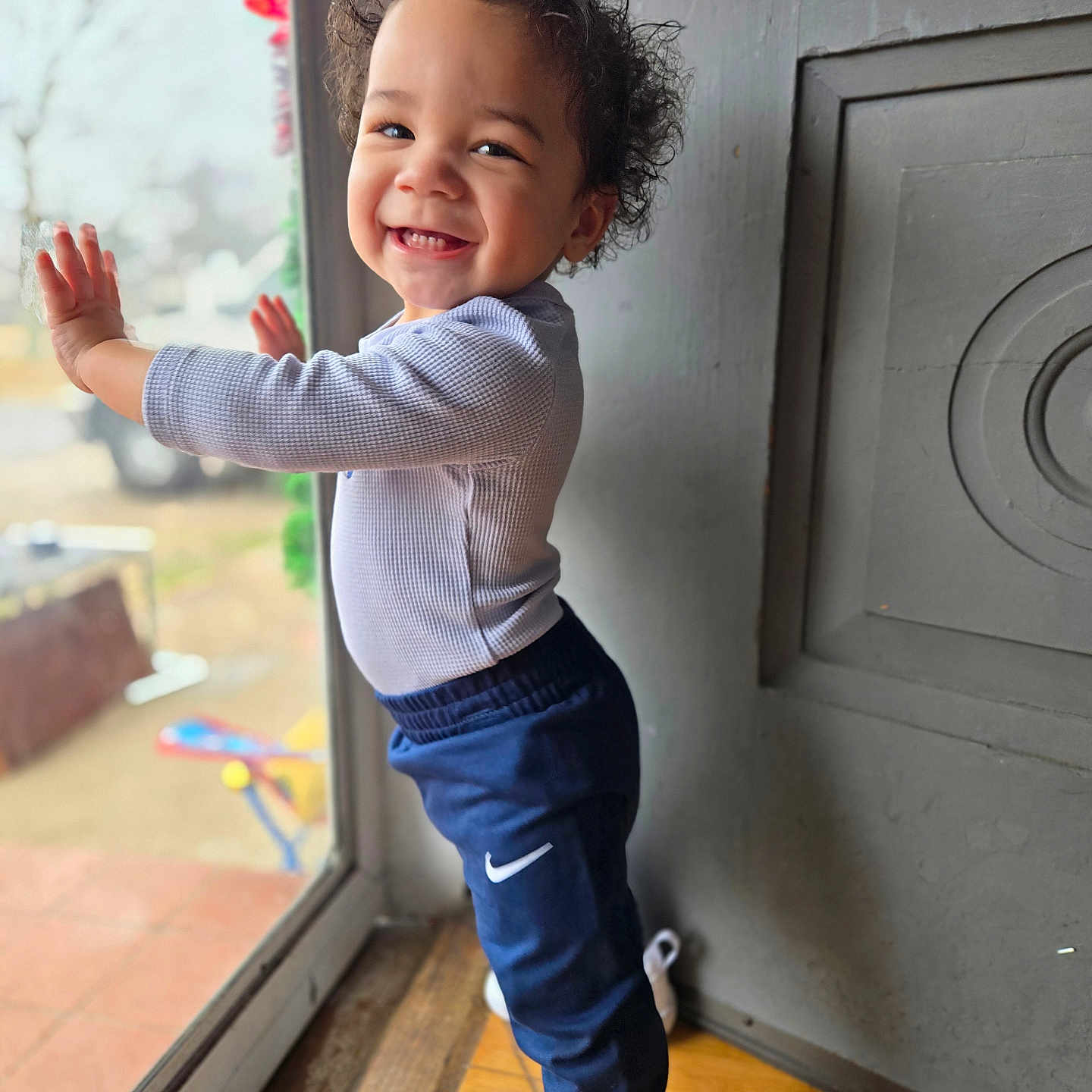 Demondre is registered to the contest to win money with this photo: casual_clothing, child, curly_hair, daylight, door, glass, happy, indoor, long_sleeve_shirt, natural_light, pants, person, pose, smiling, sneakers, standing, toddler, window, wooden_floor, young_child