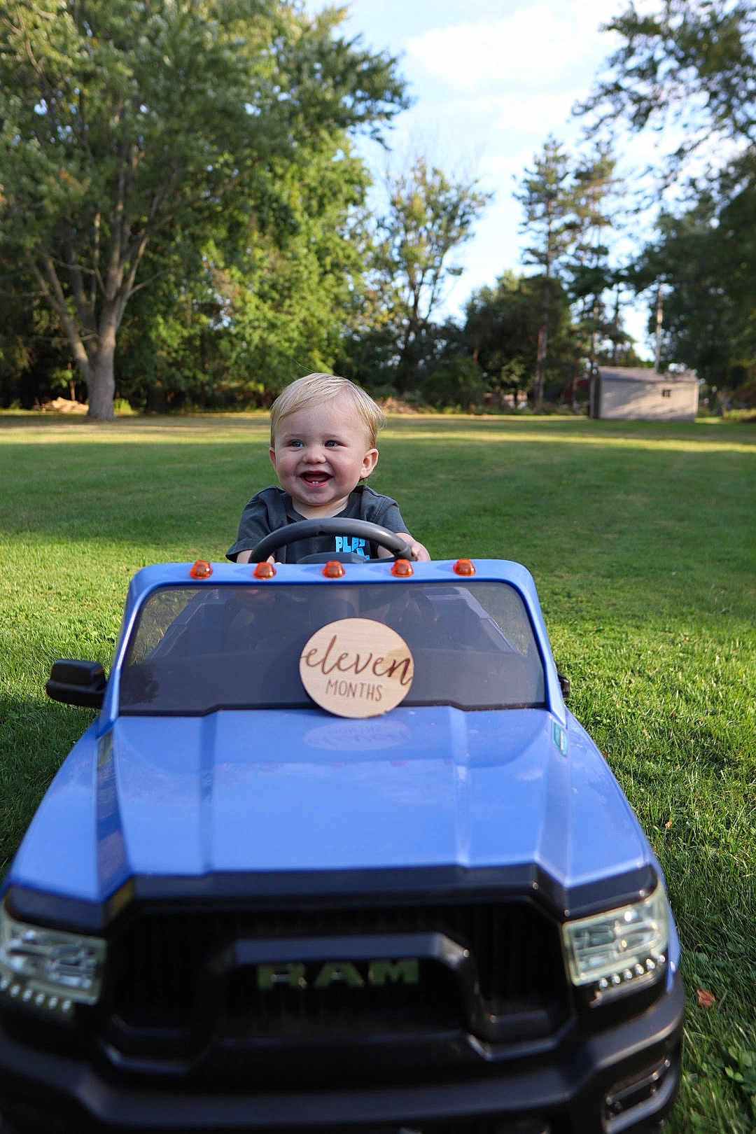Zane joined the competition — help win amazing prizes! baby, blonde_hair, blue_car, child, daylight, front_view, grass, green_lawn, happy, infant, nature, outdoor, park, play, smiling, steering_wheel, sunlight, toy_car, trees, vehicle