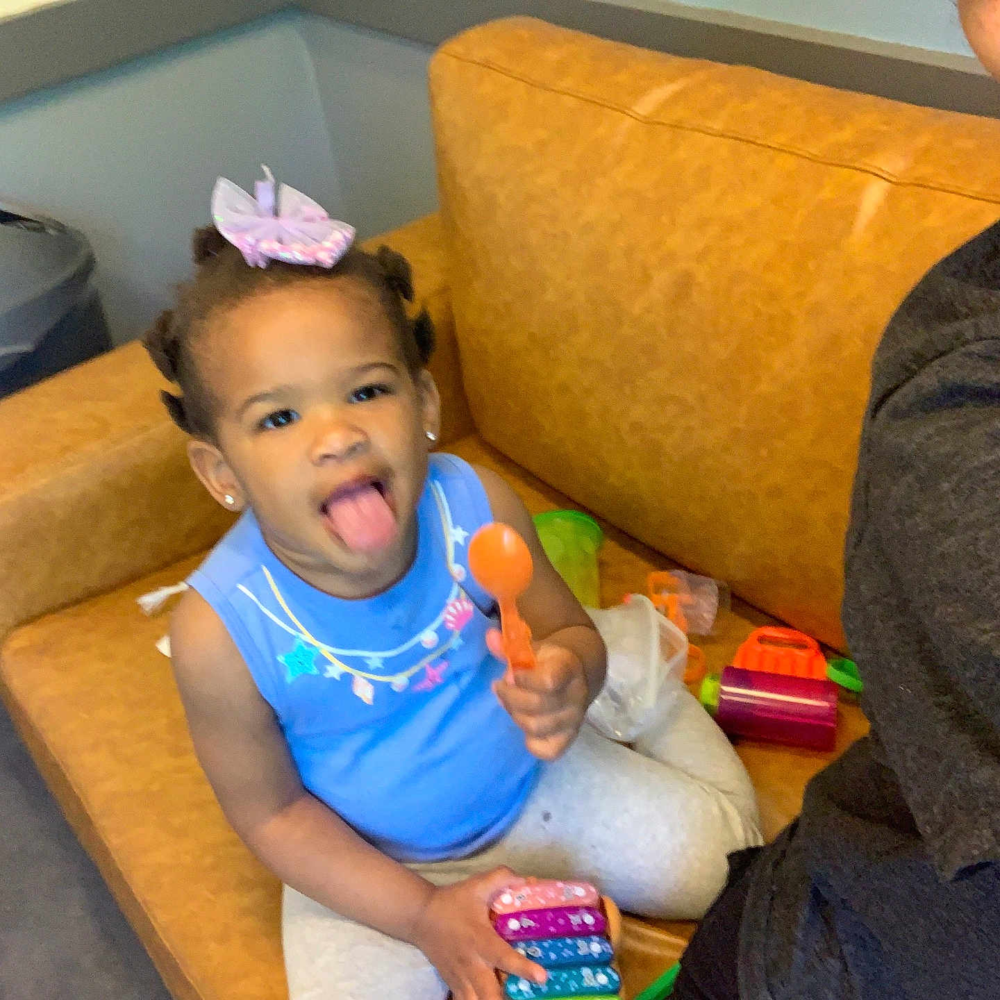 Liliana is registered to the contest to win money with this photo: toddler, child, toy, couch, indoor, playful, blue_shirt, person, smile, tongue_out, hair_bow, earrings, plastic_toys, casual, home, carpet, trash_can, happy, family, fun