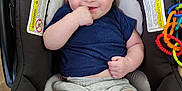 Jose is registered to the contest to win money with this photo: baby, child, car_seat, blue_shirt, gray_shorts, smiling, finger_in_mouth, toys, colorful, floor, indoors, infant, happy, cute, person, seatbelt, playful, comfort, young, portrait