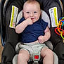 baby, child, car_seat, blue_shirt, gray_shorts, smiling, finger_in_mouth, toys, colorful, floor, indoors, infant, happy, cute, person, seatbelt, playful, comfort, young, portrait