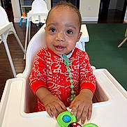 Tylan joined the competition — help win amazing prizes! toddler, child, high_chair, toy, red_clothing, indoor, wooden_floor, smiling, person, baby, closeup, playful, furniture, seat, cute, patterned_clothing, young_child, hands, face, expression