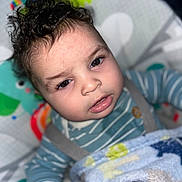 Royal is registered to the contest to win money with this photo: baby, child, curly_hair, striped_clothing, blue_blanket, infant, face, hand, baby_seat, colorful_background, cute, indoors, portrait, young_child, soft_texture, warm_clothing, sleepy, close_up, expression, comfort