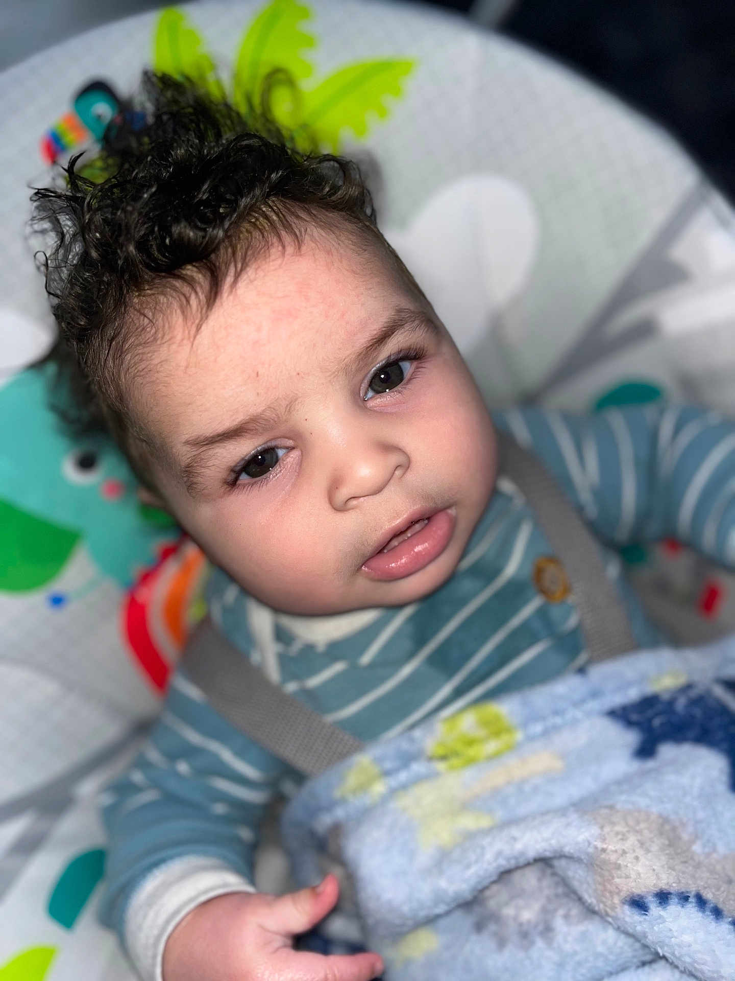 Royal is registered to the contest to win money with this photo: baby, child, curly_hair, striped_clothing, blue_blanket, infant, face, hand, baby_seat, colorful_background, cute, indoors, portrait, young_child, soft_texture, warm_clothing, sleepy, close_up, expression, comfort
