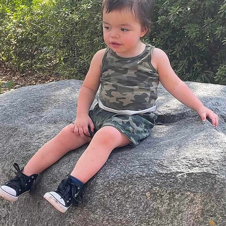 Liam joined the competition — help win amazing prizes! black_shoes, camouflage_clothing, casual_wear, child, curious, daylight, fence, greenery, nature, outdoor, person, relaxing, rock, shoes, shorts, sitting, sunlight, tank_top, toddler, young