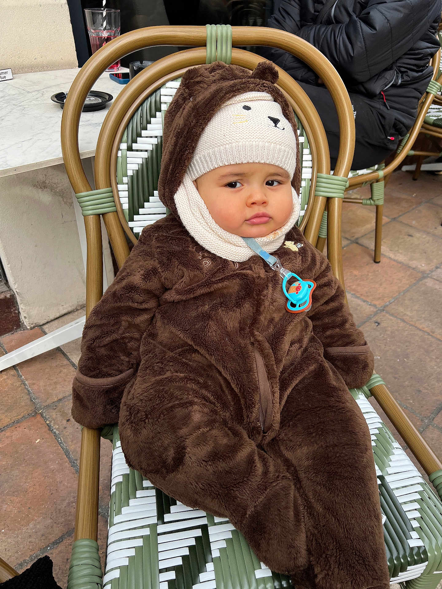 Youssef Chawachi participe au concours pour gagner de l'argent avec cette photo : baby, child, infant, bear_suit, knitted_hat, pacifier, chair, woven_chair, outdoor, pavement, brown_clothing, cute, expression, winter_clothing, seated, person, face, cold_weather, wearing, costume