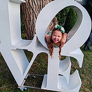 Adèlya a rejoint le concours — aidez-le/la à gagner de superbes lots ! child, girl, flower_crown, love_sculpture, outdoor, grass, tree, smiling, playful, portrait, greenery, summer, happy, casual_clothing, nature, daylight, grass_field, art_installation, young, fun