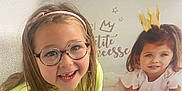 Adèlya participe au concours pour gagner de l'argent avec cette photo : child, girl, glasses, headband, smiling, sitting, indoor, poster, crown, princess, text, floor, wooden_floor, jeans, yellow_sweater, chair, window_blinds, wall, happy, portrait