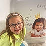 child, girl, glasses, headband, smiling, sitting, indoor, poster, crown, princess, text, floor, wooden_floor, jeans, yellow_sweater, chair, window_blinds, wall, happy, portrait
