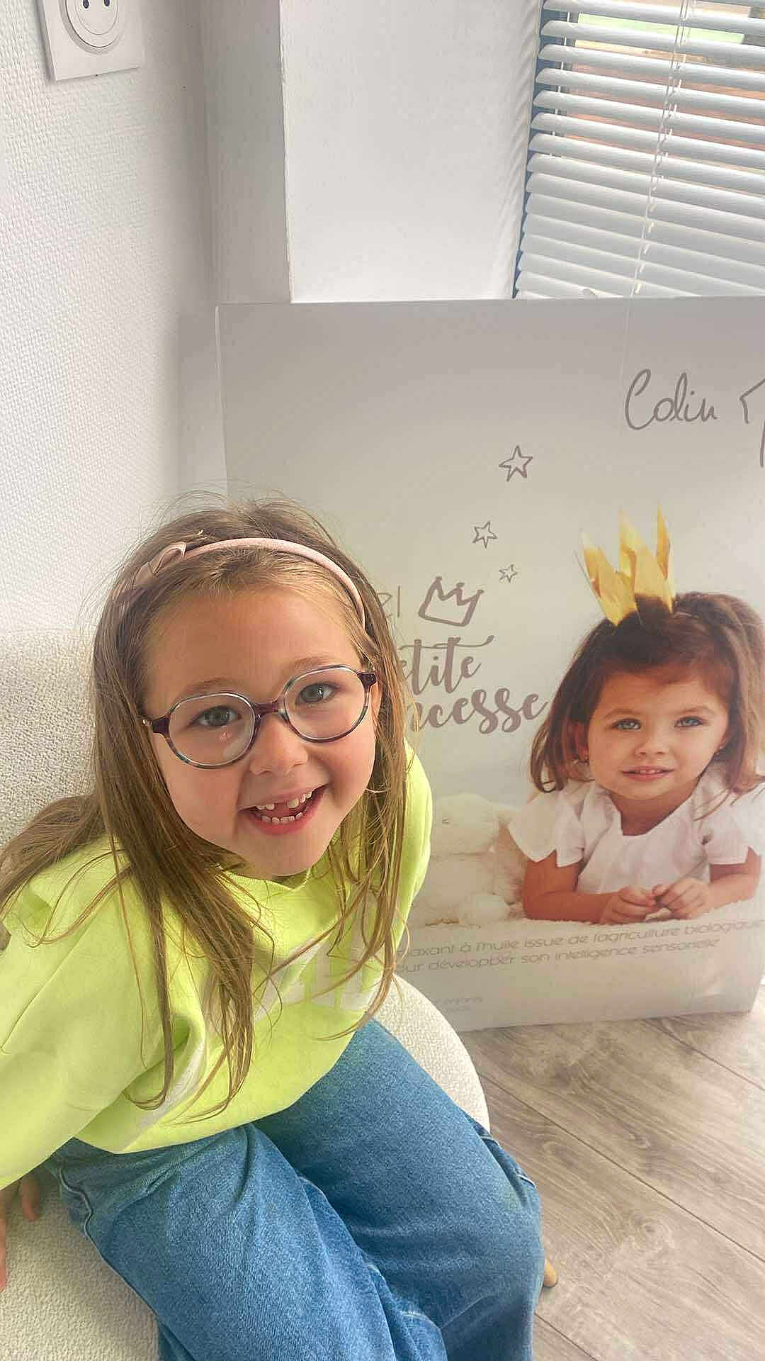 Adèlya participe au concours pour gagner de l'argent avec cette photo : child, girl, glasses, headband, smiling, sitting, indoor, poster, crown, princess, text, floor, wooden_floor, jeans, yellow_sweater, chair, window_blinds, wall, happy, portrait