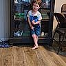barefoot, blue_outfit, boy, cabinet, chair, child, clothing, family_photo, glass_door, home, indoors, living_room, portrait, shoes, shorts, smiling, standing, table, toddler, wooden_floor