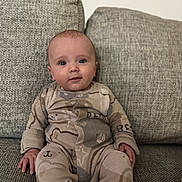 Lorenzo joined the competition — help win amazing prizes! baby, child, onesie, bear_pattern, couch, gray_couch, seated, indoors, comfortable, cute, infant, portrait, fabric_texture, clothing, human, face, hands, expression, soft_lighting, casual