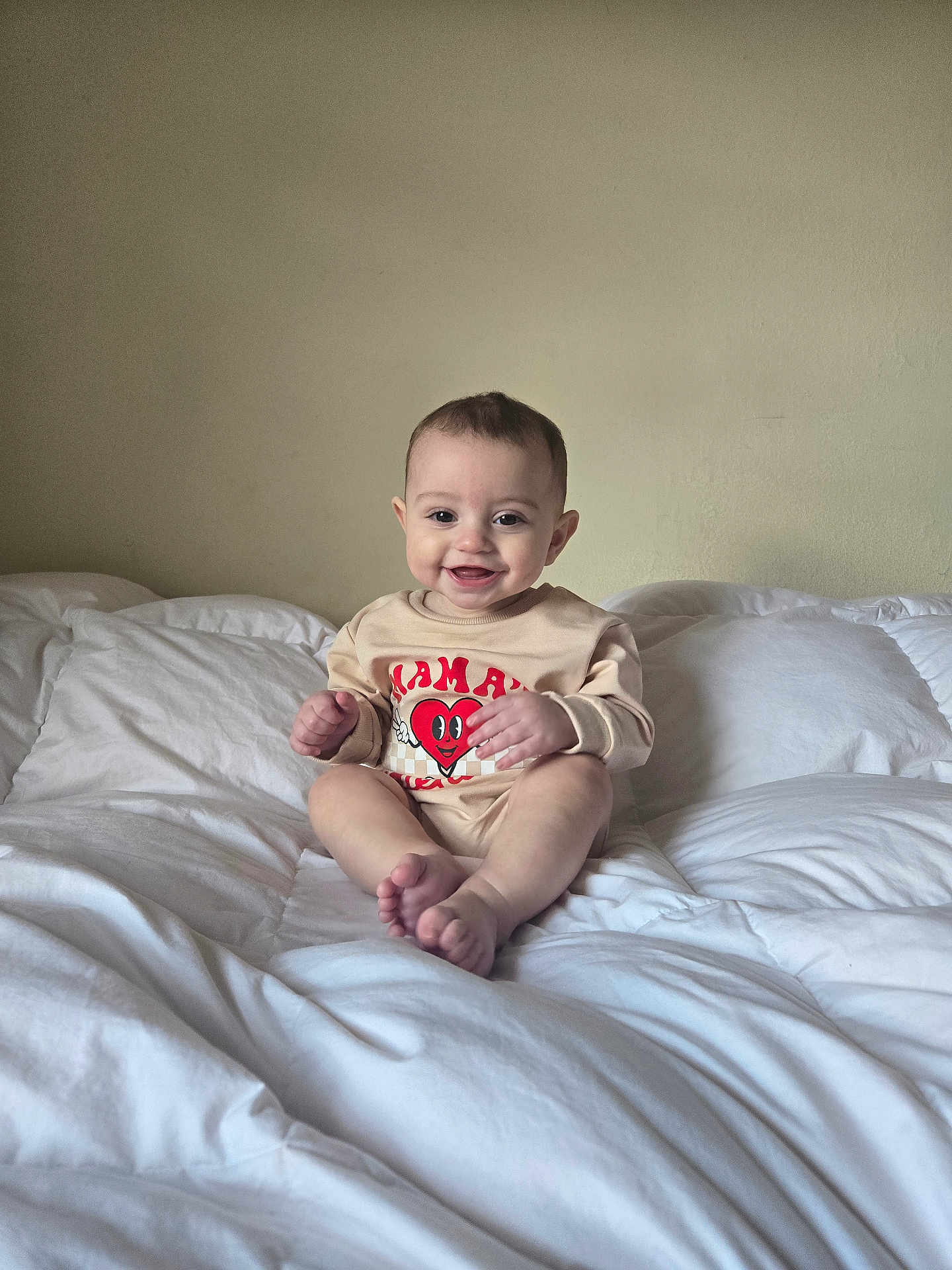 Liam is registered to the contest to win money with this photo: baby, smiling, infant, bed, white_bedding, indoors, child, clothing, happy, portrait, sitting, cute, one_person, wall, room, casual, young_child, feet, hands, head