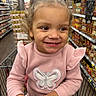 toddler, child, smiling, shopping_cart, grocery_store, aisle, canned_goods, curly_hair, pink_sweater, butterfly, happy, indoor, person, retail, food, shopping, cute, portrait, young_child, clothing