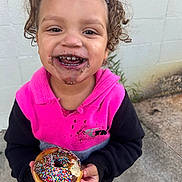 Kaiden is registered to the contest to win money with this photo: child, smiling, donut, sprinkles, food, chocolate, curly_hair, hoodie, pink_clothing, black_clothing, face, happy, outdoor, concrete, wall, holding, messy, cute, portrait, young