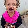 child, smiling, donut, sprinkles, food, chocolate, curly_hair, hoodie, pink_clothing, black_clothing, face, happy, outdoor, concrete, wall, holding, messy, cute, portrait, young