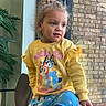 child, toddler, sitting, yellow_sweatshirt, blue_pants, cartoon_characters, curly_hair, indoor, chair, plant, window, brick_wall, natural_light, casual_clothing, happy, portrait, person, smiling, home, cozy
