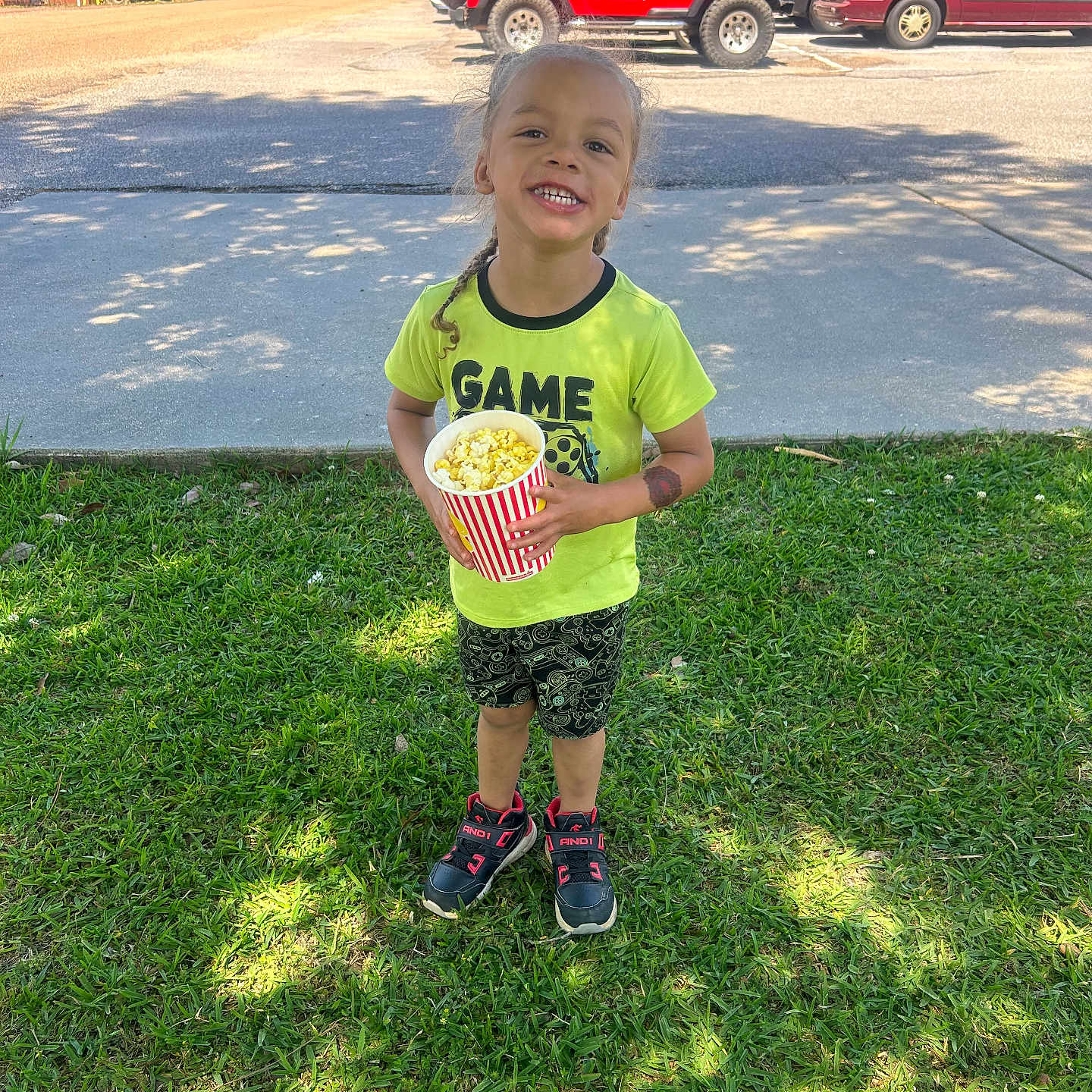 Jadev is registered to the contest to win money with this photo: child, popcorn, grass, outdoor, smiling, toddler, green_shirt, shorts, sneakers, sunlight, tree, vehicle, parking_lot, sidewalk, happy, playful, daytime, casual_clothing, person, nature