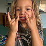 child, curly_hair, smiling, hand_gesture, tshirt, indoor, television, carpet, furniture, person, playful, young, casual_clothing, portrait, happy, hands, living_room, home, cute, expression