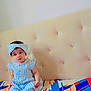 Minha is registered to the contest to win money with this photo: baby, child, blue_striped_dress, headband, sitting, colorful_blanket, beige_headboard, indoors, curious_expression, cute, portrait, young_child, patterned_blanket, furniture, soft_lighting, person, clothing, headwear, infant, room