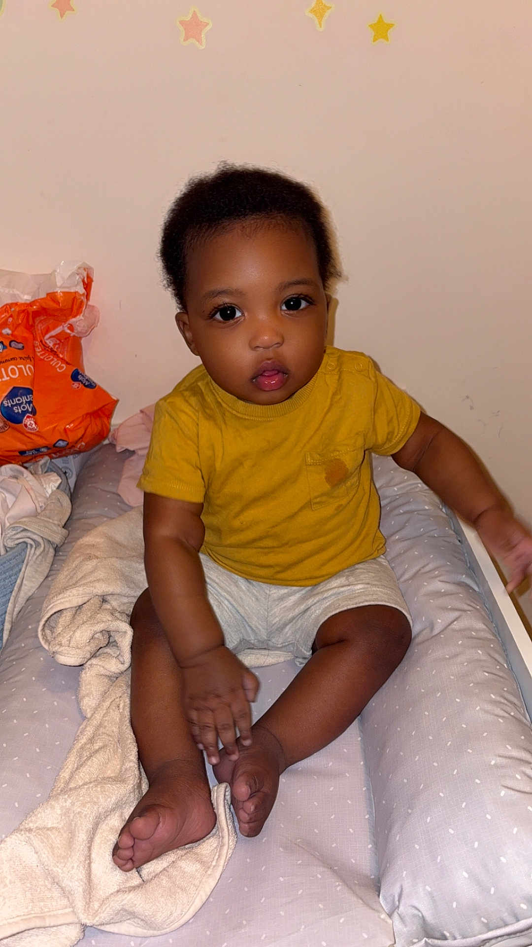 Adam participe au concours pour gagner de l'argent avec cette photo : toddler, child, baby, sitting, mustard_shirt, shorts, cushioned_changing_pad, blanket, soft_toys, indoor, wall_stickers, star_stickers, curious_eyes, dark_skin, short_hair, hand, feet, clothing, baby_essentials, room