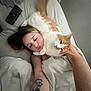 person, cat, bed, blanket, tattoo, pillow, remote_control, white_sheets, orange_cat, lying_down, affection, selfie, indoor, relaxing, cute, pet, sleepy, cozy, human_face, animal