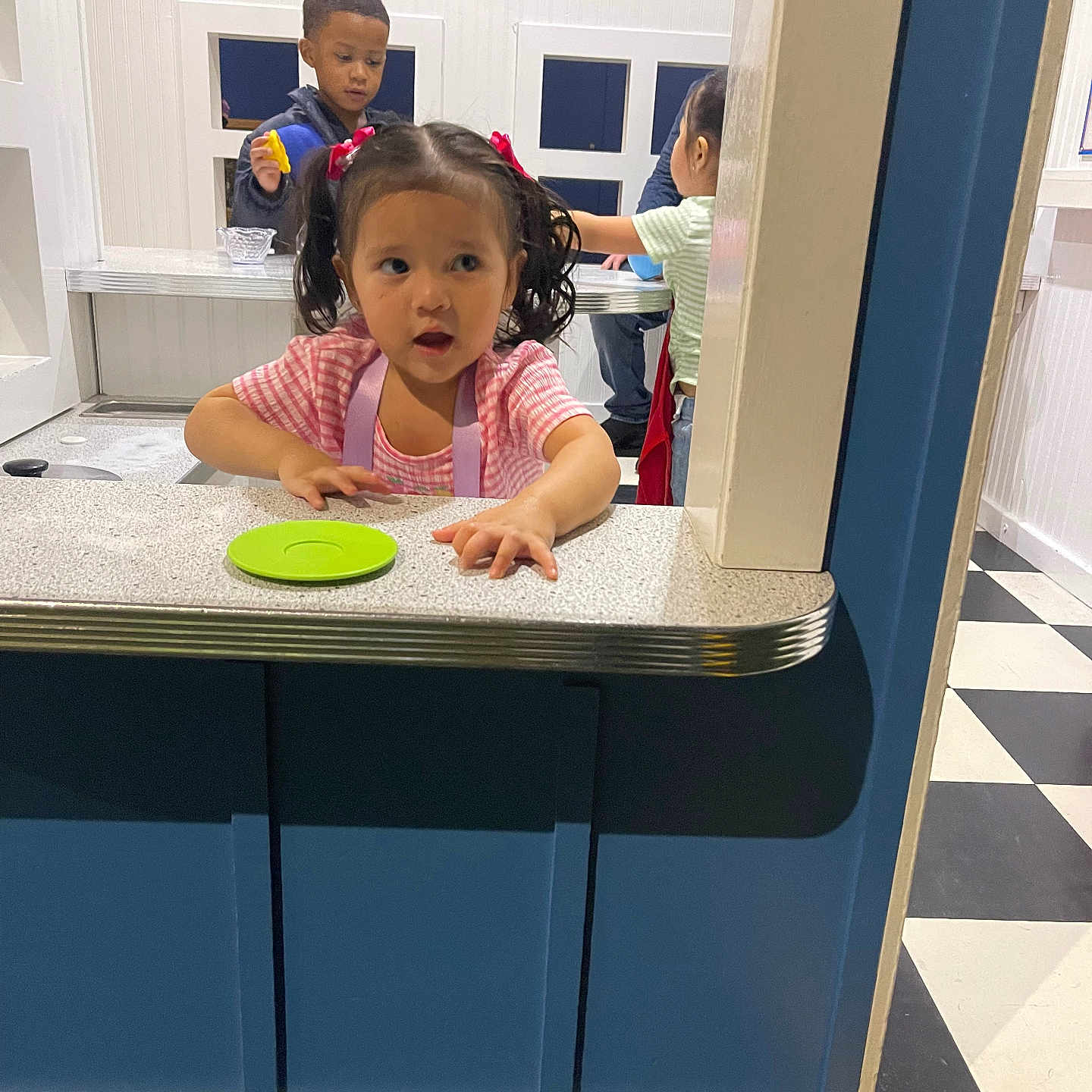 Leiliany is registered to the contest to win money with this photo: apron, blue_wall, casual_clothing, checkered_floor, child, countertop, curious, floor, girl, indoor, interaction, kids, pigtails, play, playroom, table, toy, white_wall, window, young