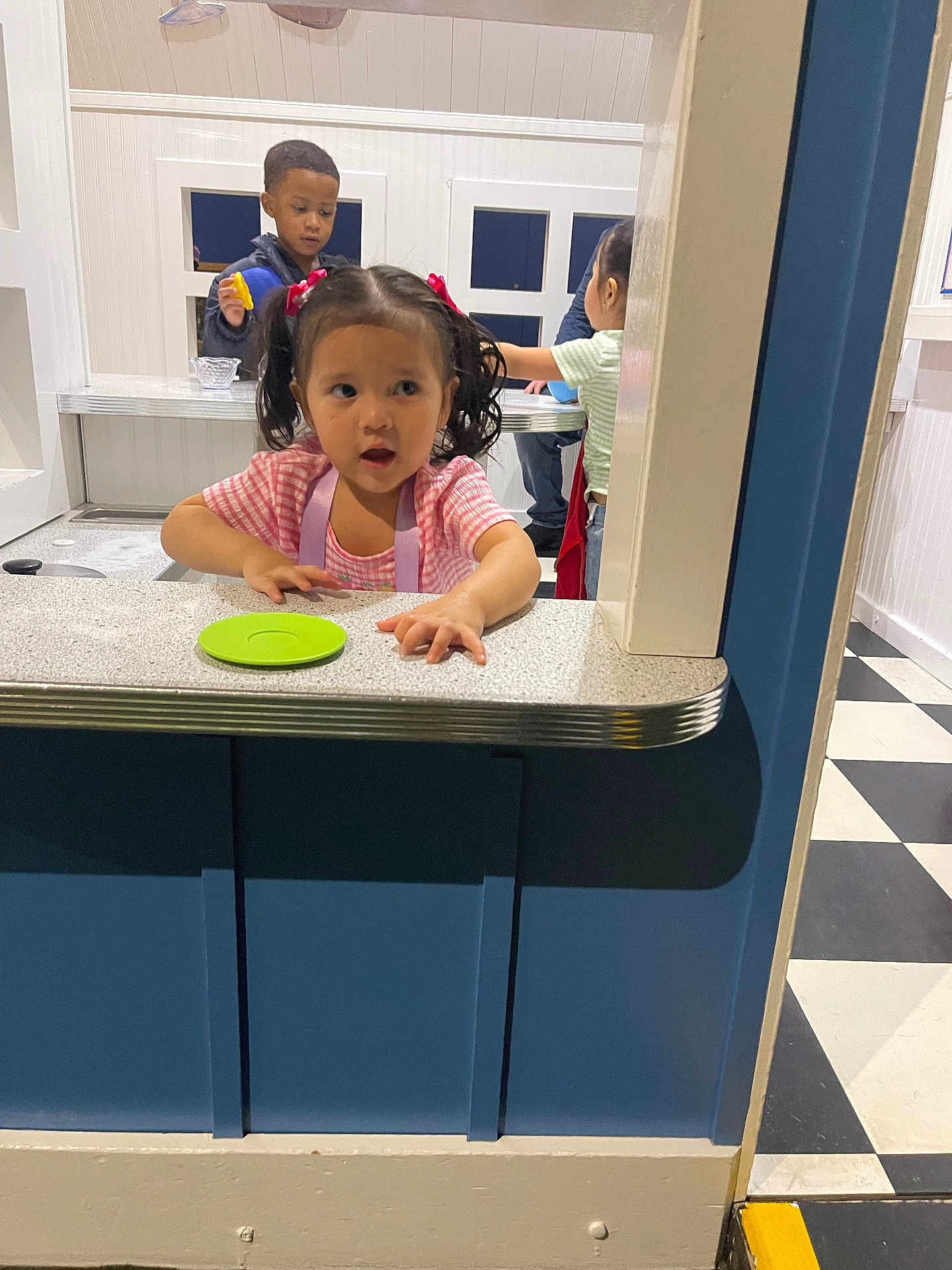 Leiliany is registered to the contest to win money with this photo: child, girl, pigtails, countertop, indoor, playroom, kids, curious, apron, table, window, floor, checkered_floor, white_wall, blue_wall, toy, play, interaction, young, casual_clothing