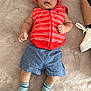 baby, infant, child, hooded_vest, red_clothing, blue_shorts, socks, blanket, floor, lotion_bottle, person, footwear, white_sneaker, indoor, soft_texture, casual_clothing, lying_down, curious_expression, small_hands, cute