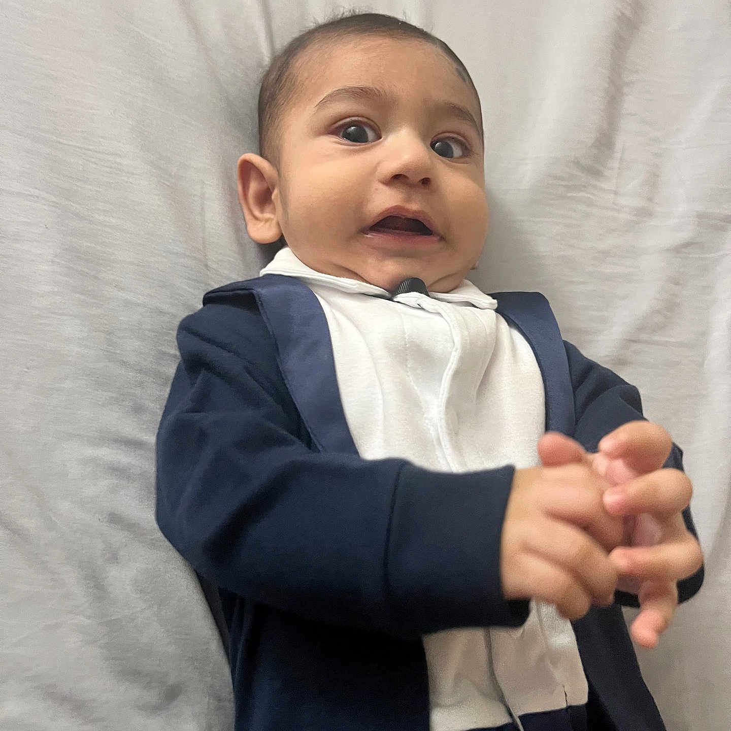 Iyad is registered to the contest to win money with this photo: baby, bed, blanket, casual, child, clothing, cute, expression, face, funny, hands, indoor, infant, laying, onesie, person, portrait, surprised, tuxedo, young
