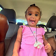 Melody is registered to the contest to win money with this photo: child, toddler, pink_dress, car_seat, pacifier, necklace, inside_car, playful, tongue_out, smiling, person, seat, window, adult_arm, curly_hair, sunlight, casual, happy, portrait, fun