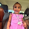 child, toddler, pink_dress, car_seat, pacifier, necklace, inside_car, playful, tongue_out, smiling, person, seat, window, adult_arm, curly_hair, sunlight, casual, happy, portrait, fun