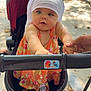 baby, child, stroller, hand, outdoor, park, bench, sunlight, floral_dress, head_wrap, person, infant, holding_hand, toy, wheel, shadow, pavement, summer, expression, curious