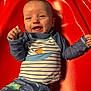baby, child, smiling, happy, red_chair, striped_clothing, patterned_clothing, indoor, infant, cute, joyful, portrait, person, sitting, playful, colorful, raised_arms, young_child, face, clothing