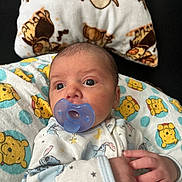Asher is registered to the contest to win money with this photo: baby, newborn, infant, child, portrait, pacifier, lying_down, blanket, pillow, onesie, winnie_the_pooh, stuffed_animal_pattern, blue, yellow, indoor, cozy, wide_eyed, adult_hand, close_up, family