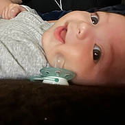 Bryce is registered to the contest to win money with this photo: baby, infant, pacifier, face, eyes, clothing, gray_outfit, lying_down, closeup, skin, cute, child, newborn, indoors, portrait, head, expression, small_hand, dark_background, soft_texture
