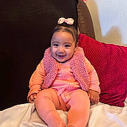 Carolin is registered to the contest to win money with this photo: baby, child, smile, pink_clothing, bow, vest, couch, cushion, happy, indoor, portrait, infant, sitting, cozy, furniture, cute, person, headwear, soft_texture, warm_colors