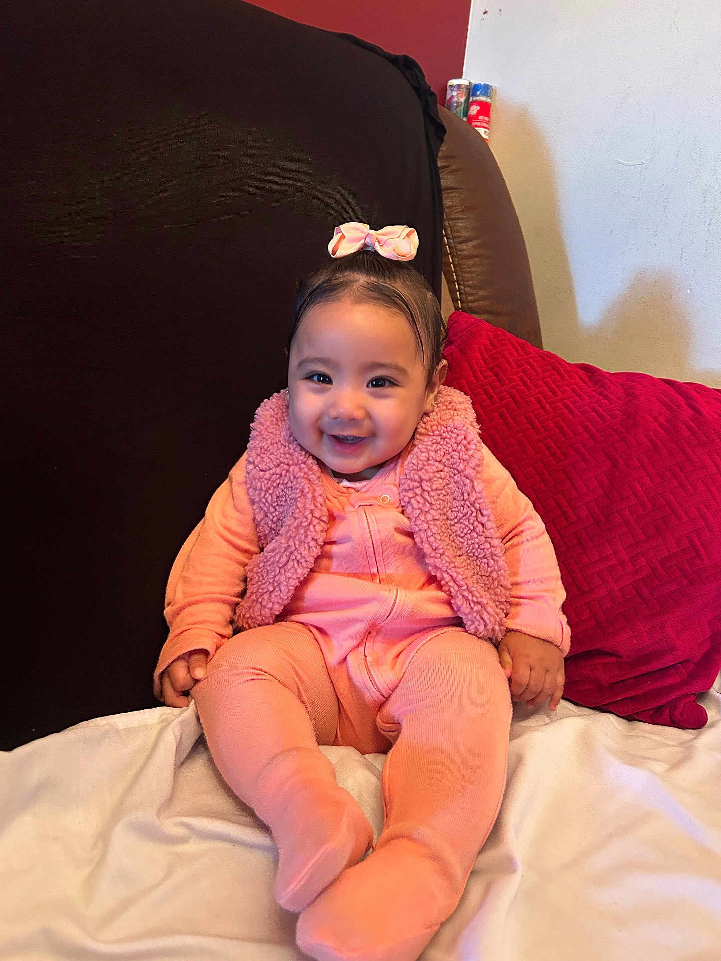 Carolin is registered to the contest to win money with this photo: baby, child, smile, pink_clothing, bow, vest, couch, cushion, happy, indoor, portrait, infant, sitting, cozy, furniture, cute, person, headwear, soft_texture, warm_colors