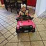 toddler, child, toy_car, pink, white_bow, indoor, tile_floor, table, chairs, steering_wheel, person, floor, play, cute, baby, vehicle, seatbelt, furniture, home, girl