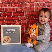 Lenora joined the competition — help win amazing prizes! baby, child, teddy_bear, brick_wall, sign, milestone, smile, sitting, plush_toy, soft_surface, indoor, cute, portrait, happy, clothing, earrings, blue, orange, frame, text
