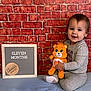 baby, child, teddy_bear, brick_wall, sign, milestone, smile, sitting, plush_toy, soft_surface, indoor, cute, portrait, happy, clothing, earrings, blue, orange, frame, text