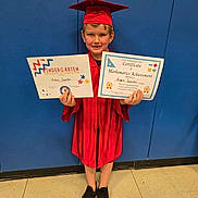 Aidan joined the competition — help win amazing prizes! child, young_student, graduation, graduation_cap, red_gown, certificate, achievement, kindergarten, mathematics, school, portrait, standing, smiling, indoor, blue_background, school_floor, award, education, shoes, proud