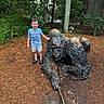 child, boy, smiling, outdoor, garden, gorilla, statue, animal_statue, wildlife, nature, trees, plants, greenery, wooden_fence, mulch, playground, zoo, posing, portrait, adventure