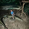 child, crocodile, reptile, jungle, outdoor, wildlife, animal, adventure, smiling, sitting, blue_shirt, shorts, shoes, tree, roots, wooden_sign, sand, exhibit, nature, portrait