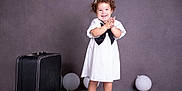 Meghan joined the competition — help win amazing prizes! toddler, child, smiling, white_dress, black_bow, curly_hair, sandals, balloon, suitcase, studio, floor_reflection, portrait, happy, clapping, indoor, photography, cute, standing, background, fashion
